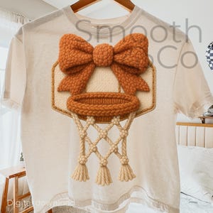 May include: Cream-colored t-shirt featuring a basketball hoop design with a bow. The hoop, net, and bow are in shades of orange and beige, creating a textured, handmade appearance. The text "Smooth Co" is visible on the shirt.