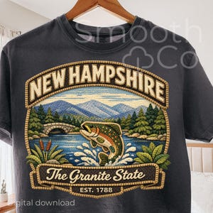May include: Dark gray t-shirt featuring a detailed embroidered design of New Hampshire's landscape. The design includes a jumping fish, mountains, a bridge, and the text "New Hampshire" and "The Granite State". The shirt is hanging on a wooden hanger.