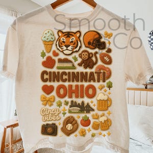 Op de afbeelding: Crèmekleurig T-shirt met een kleurrijk grafisch ontwerp met illustraties met het thema Cincinnati, Ohio. Het ontwerp bevat een tijger, een footballhelm, een aap en de woorden "Cincinnati Ohio" en "Cincy vibes".