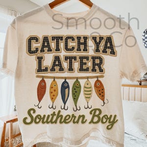 Op de afbeelding: Crèmekleurig T-shirt met de tekst "CATCH YA LATER" in een ruitpatroon. Daaronder hangen zes kleurrijke vissenkunstaasjes aan een lijn, met de woorden "Southern Boy" eronder.