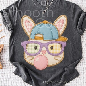 May include: A dark gray t-shirt featuring a cartoon bunny face with a blue baseball cap, purple glasses, and a pink bubble gum bubble. The bunny's ears are beige with pink gingham lining. The glasses have green and yellow gingham lenses.