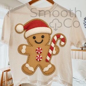 May include: Cream t-shirt with a gingerbread man design. The gingerbread man wears a red Santa hat and holds a red and white candy cane. The design includes two red and white candies and is outlined with brown stitching. The text "Smooth Co" is visible.