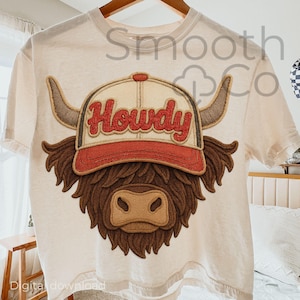 May include: A cream-coloured t-shirt with a brown Highland cow design. The cow wears a red and tan baseball cap with the word "Howdy" in red script. The design includes horns and shaggy fur.