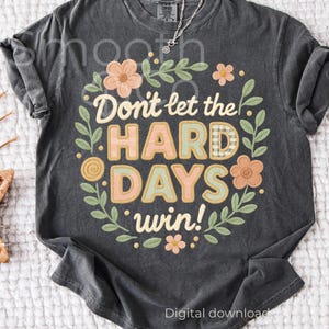 Op de afbeelding: Donkergrijs t-shirt met de tekst "Don't let the HARD DAYS win!" in een bloemenkrans. Het ontwerp gebruikt pastelkleuren en een ruitpatroon. De woorden zijn omgeven door bloemen en bladeren.