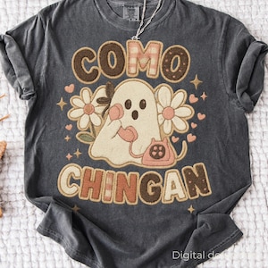Könnte beinhalten: Dunkelgraues T-Shirt mit einem Grafikdesign, das einen Geist mit einem rosa Telefon zeigt, umgeben von Blumen und Herzen. Der Text "COMO CHINGAN" ist oberhalb und unterhalb des Geistes in braunen Buchstaben dargestellt.