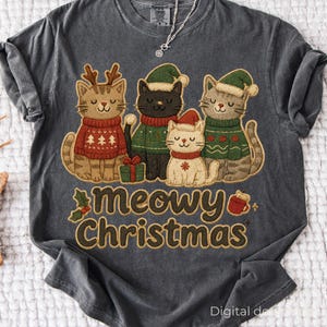 May include: Dark gray t-shirt featuring a festive design with four cartoon cats wearing Christmas sweaters and hats. The phrase "meowy Christmas" is printed in a decorative font below the cats. A small silver necklace is visible.
