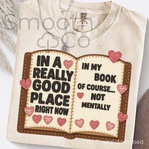 Puede incluir: Camiseta color crema con un diseño de libro abierto. El texto en el libro dice: "IN A REALLY GOOD PLACE RIGHT NOW" y "IN MY BOOK OF COURSE... NOT MENTALLY." Acentos de corazón rosa cosidos alrededor del libro.