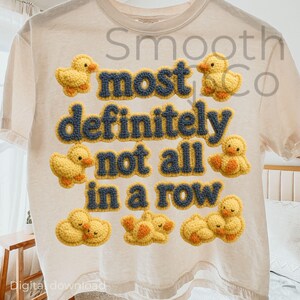 Könnte beinhalten: Cremefarbenes T-Shirt mit dem marineblauen Schriftzug "most definitely not all in a row" und gelben Häkelenten-Akzenten. Das Shirt hat eine lockere Passform und kurze Ärmel. Der Text "Smooth Co" ist oben sichtbar.