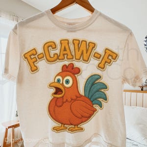 Pode incluir: Camiseta creme com um desenho de galinha de desenho animado em laranja, vermelho e azul. O texto "F-CAW-F" está acima da galinha em uma fonte amarela e marrom. A camiseta está pendurada em um cabide de madeira.