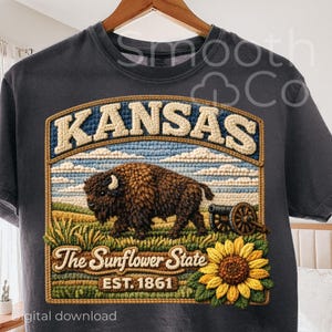 May include: Dark gray t-shirt featuring a detailed embroidered design. The design includes the word "KANSAS" in cream-colored letters, a brown bison, and a sunflower. The text "The Sunflower State EST. 1861" is also present.