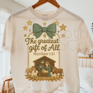 May include: Cream-coloured t-shirt with the text "The greatest gift of All" and Matthew 1:21. The design includes a green bow, gold stars, and a nativity scene with figures and animals. A casual, religious-themed tee.