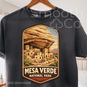 May include: Dark gray t-shirt featuring a Mesa Verde National Park design. The graphic depicts a detailed illustration of the park's cliff dwellings, with the text "MESA VERDE NATIONAL PARK" below. The design is in shades of brown, beige, and blue.
