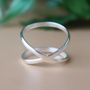 May include: A silver-colored ring with an X-shaped design. The ring features two bands that intersect in the center, creating a unique and modern look. The ring is set against a neutral background, highlighting its sleek and minimalist style.