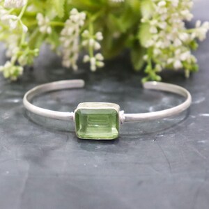 May include: A silver cuff bracelet featuring a rectangular, light green gemstone. The bracelet has a simple, elegant design, with the gemstone set in a square silver bezel. The background includes blurred white flowers.
