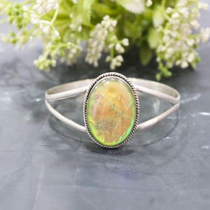 May include: A silver cuff bracelet with an oval-shaped, iridescent gemstone. The gemstone has a mix of yellow, green, and orange hues, and is framed by a twisted silver border. The bracelet is displayed on a dark surface with white flowers in the background.