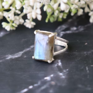 May include: A silver ring with a rectangular labradorite gemstone. The stone has iridescent blue and grey tones. The ring has a simple, modern design with a split shank and four corner prongs holding the stone.