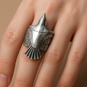 May include: A silver-colored ring in the shape of a bird with outstretched wings. The ring is detailed with feathers and a pointed beak. The ring is worn on a finger against a neutral background.