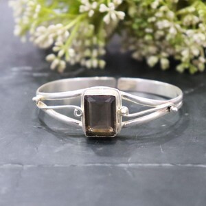 May include: A silver-toned bracelet with a rectangular, smoky quartz gemstone at its center. The bracelet features a split band design with decorative accents. The background includes greenery and a dark surface.