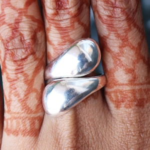 Peut inclure: Une bague argentée brillante avec un design unique et fluide. La bague présente deux éléments en forme de goutte d'eau qui s'enroulent autour du doigt. Le métal poli reflète la lumière, créant un aspect lumineux et attrayant. Bijoux.