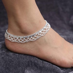 May include: A silver-toned anklet featuring a delicate chain design with a layered, scalloped lattice pattern. The jewelry piece consists of a smooth upper chain and a decorative, intricate lower section that creates a repeating wave-like structure.