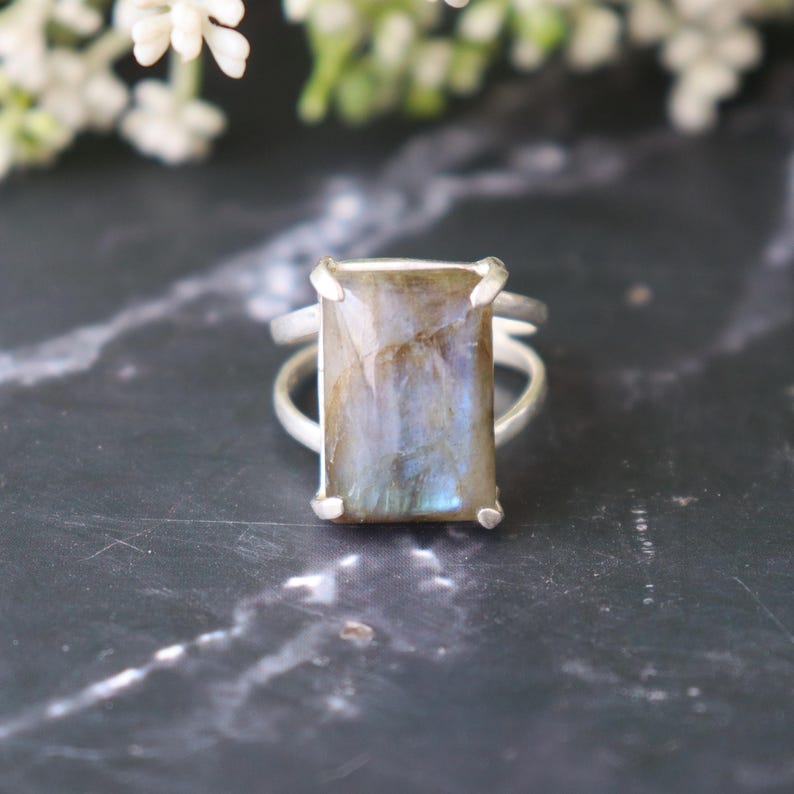 May include: A silver ring with a rectangular labradorite gemstone. The stone has blue, grey, and brown tones. The ring's band is plain, with the stone secured by four prongs. The ring is a statement piece.