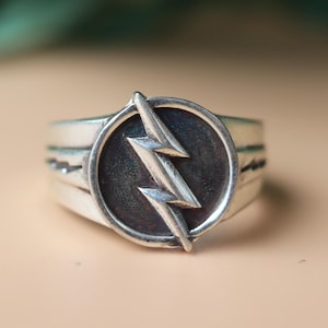 Könnte beinhalten: Ein silberner Ring mit einem kreisförmigen Emblem, das einen Blitz zeigt. Der Ring hat ein strukturiertes Band mit drei horizontalen Linien. Der Blitz hat eine kontrastierende dunkle Farbe vor einem helleren silbernen Hintergrund.