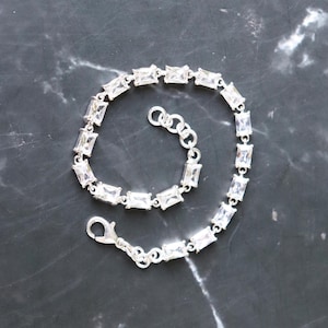 May include: A silver bracelet with rectangular, clear gemstones. The bracelet is laid out on a dark, marbled surface. The clasp is visible, and the bracelet appears to be made of silver.
