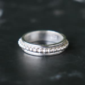 Puede incluir: Un anillo de plata con un diseño de cuentas alrededor de la banda. El anillo tiene un acabado liso y pulido y un estilo simple y elegante. El anillo es probablemente de plata de ley.