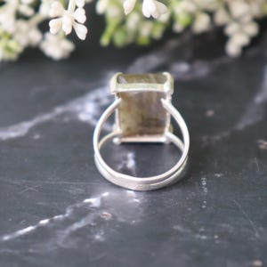 May include: A silver ring featuring a rectangular, iridescent gemstone. The ring has a double-band design and a simple setting. The gemstone displays green and brown hues. The ring is presented on a dark, marble-effect surface.