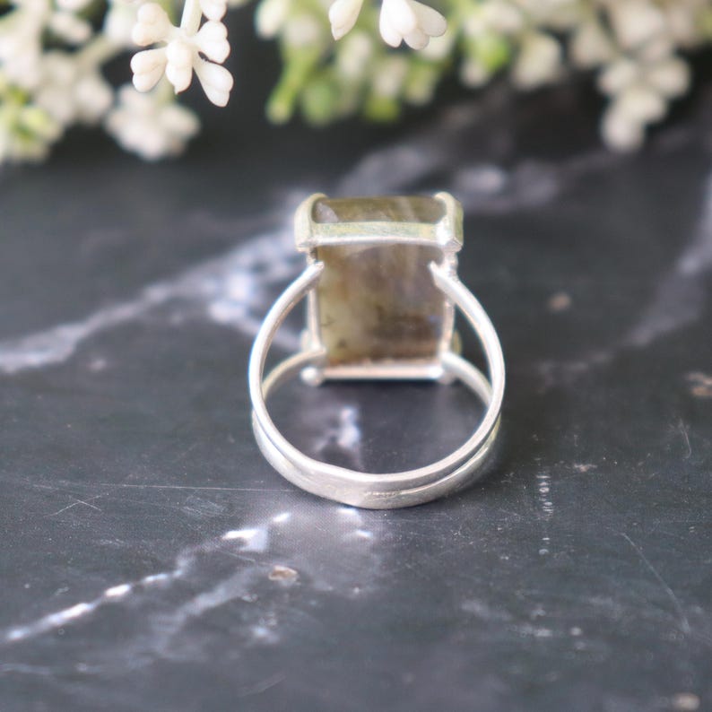 May include: A silver ring featuring a rectangular, iridescent gemstone. The ring has a double-band design and a simple setting. The gemstone displays green and brown hues. The ring is presented on a dark, marble-effect surface.