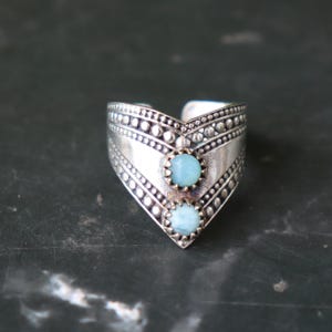 May include: A silver-toned ring with a chevron design, featuring two light blue gemstones. The ring has a textured surface with dotted and linear patterns, creating a vintage or bohemian aesthetic.