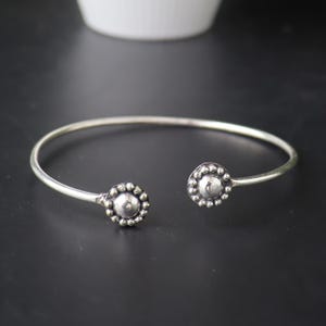 Könnte beinhalten: Ein silberfarbenes offenes Armband. Jedes Ende hat ein kreisförmiges Design mit einer zentralen Perle, die von kleineren Perlen umgeben ist. Das Armband wird auf einer dunklen Oberfläche mit einem verschwommenen weißen Objekt im Hintergrund präsentiert.