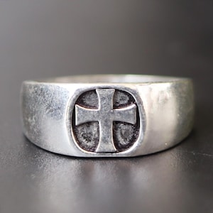 Puede incluir: Un anillo de plata con un diseño de cruz. El anillo tiene una banda ancha y una superficie ligeramente texturizada, con la cruz dentro de un óvalo. La cruz tiene una forma clásica y simétrica, y el anillo tiene una apariencia vintage o antigua.