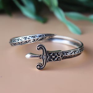 Puede incluir: Un anillo plateado con un diseño abierto, con un adorno en forma de espada. El anillo tiene patrones intrincados y detallados a lo largo de la banda y en la empuñadura de la espada. El anillo se muestra sobre una superficie de tono neutro.