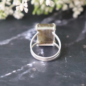 May include: A silver ring with a rectangular, iridescent gemstone. The ring has a double-band design and a simple setting. The gemstone shows green and brown hues. The ring is set against a dark, marble-effect surface.