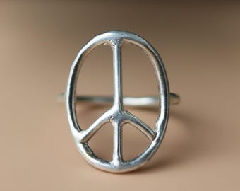 Könnte beinhalten: Ein silberfarbener Ring mit einem ovalen Friedenszeichen als Herzstück. Das Metall weist eine leicht texturierte, matte Oberfläche auf, wobei ein durchgehendes Band an die Seiten des vertikalen Friedenssymbols anschließt.