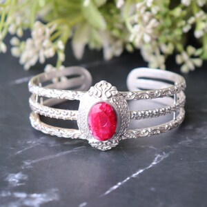 May include: A silver-toned cuff bracelet featuring three parallel bands with intricate embossed detailing. The center of the bracelet showcases a prominent, oval-shaped, faceted bright pink stone set within an ornate, decorative silver-toned frame.