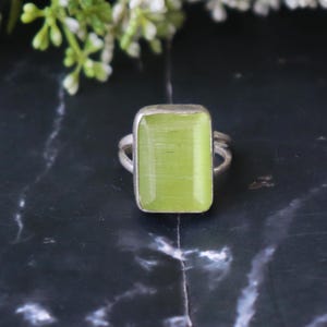 May include: A silver ring featuring a rectangular, light green gemstone. The gemstone has a smooth, polished surface and is set in a silver band. The ring's design is simple and elegant, with a focus on the gemstone's color and shape.