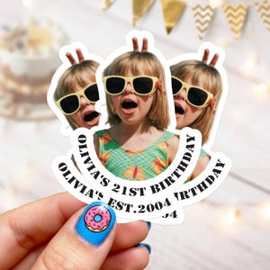 Può includere: Tre adesivi con una ragazza con occhiali da sole e un top verde floreale. Gli adesivi hanno il testo "OLIVIA'S 21ST BIRTHDAY EST. 2004" in un banner curvo. Gli adesivi sono tenuti da una mano con smalto blu e un disegno a ciambella.