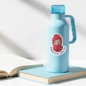 Puede incluir: Una botella de agua azul claro con un asa y tapa azules a juego. Una pegatina en la botella presenta a una persona con gorro y bufanda de punto rojos, con las palabras "FOREVER YOUNG" en un dise&ntilde;o circular. La botella descansa sobre un libro azul claro.