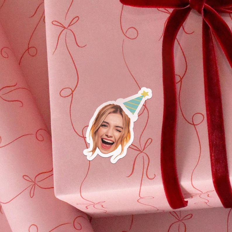 Puede incluir: Una pegatina con una persona sonriente con un gorro de fiesta. La pegatina est&aacute; colocada en una caja de regalo rosa con cintas y lazos rojos. El fondo es rosa suave con un patr&oacute;n repetido de lazos rojos.