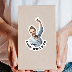 Puede incluir: Una pegatina con el texto "Look Who's 20" que rodea una foto de una persona sonriente con el brazo levantado. La pegatina est&aacute; sobre una caja marr&oacute;n claro.