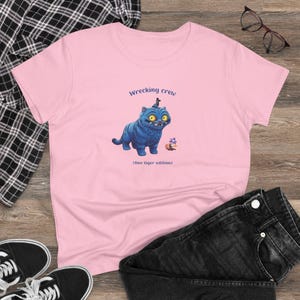 May include: A light pink t-shirt featuring a blue cartoon cat with yellow eyes and the text "Wrecking crew." Below the cat is the text "(One tiger edition)." The shirt is paired with black jeans and black sneakers.