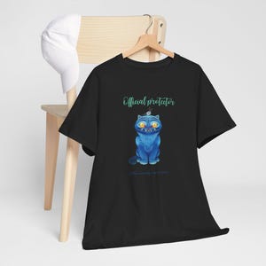 May include: Black t-shirt featuring a blue cartoon cat with yellow eyes and a crown. The text "Official protector" is above the cat, with additional text below. A white baseball cap rests on a wooden chair.