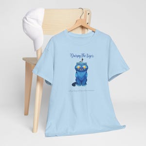 May include: A light blue t-shirt with a cartoon illustration of a blue tiger with yellow eyes and the text "Durpy the Tiger". The shirt is hanging on a wooden hanger.