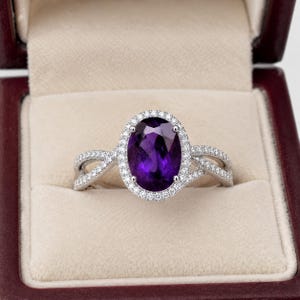 May include: A silver-toned ring featuring a large, oval-cut deep purple amethyst gemstone at the center. The stone is surrounded by a halo of small, clear pavé-set stones. The band has a split, crossover design encrusted with small clear stones.