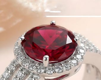 May include: Close-up of a silver ring with a large, faceted, deep red gemstone encircled by smaller, clear stones. The ring's band is also decorated with smaller, clear stones, creating a sparkling effect. The ring is set against a soft, blurred background.