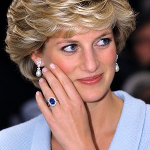 May include: An oval-shaped blue sapphire ring surrounded by a halo of small white diamonds set in a silver-toned band. The ring is worn on a finger, accompanied by matching drop earrings featuring a small diamond stud and a white pearl pendant.