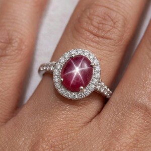 May include: A silver ring featuring a central oval-shaped cabochon star ruby with a distinct six-rayed star pattern. The ruby is surrounded by a halo of small round brilliant-cut white diamonds, with additional diamonds set along the silver band.