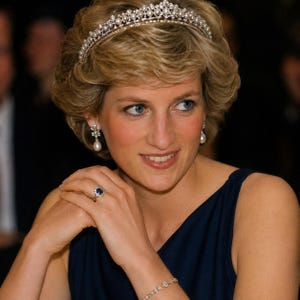 Puede incluir: Una tiara de plata con perlas y diamantes, pendientes colgantes a juego con motivos florales, un anillo con un zafiro azul central rodeado de diamantes y una delicada pulsera de diamantes. Las joyas se llevan con una prenda azul marino sin mangas.